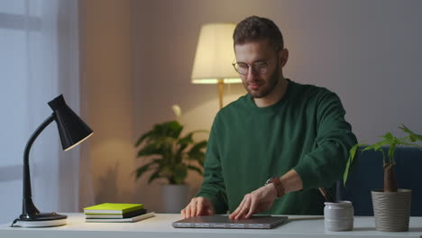 Un-Joven-Con-Gafas-Está-Usando-Una-Computadora-Portátil-Por-La-Noche-En-Casa-Abriendo-Un-Cuaderno-Y-Encendiendo-Una-Lámpara-De-Mesa-Retrato-Medio-De-Un-Hombre-Con-Gafas